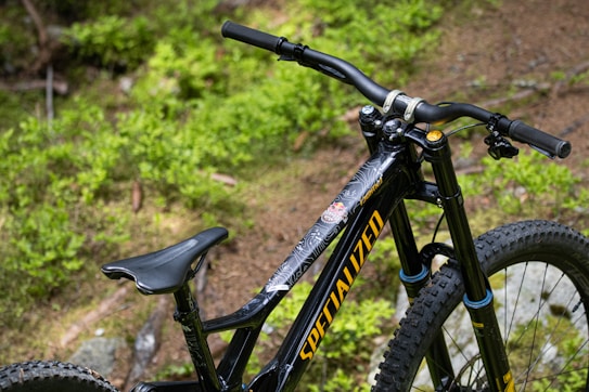 A close-up view of a black mountain bike featuring large tires and a prominent handlebar set against a backdrop of lush green foliage. The bike frame displays the brand name in bold yellow lettering.