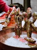 Two intricately designed brass figures are placed in a metal dish filled with white grains. The figures are adorned with red powder, commonly used in religious or ceremonial contexts. A bright garland with gold and black elements decorates the scene, resting on a terracotta object. The background includes a person dressed in red.