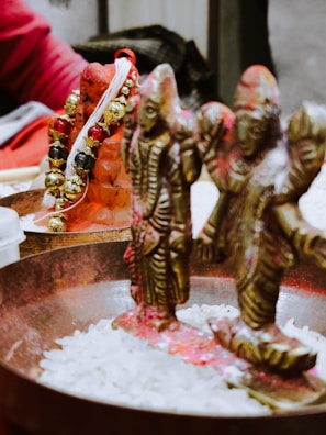 Two intricately designed brass figures are placed in a metal dish filled with white grains. The figures are adorned with red powder, commonly used in religious or ceremonial contexts. A bright garland with gold and black elements decorates the scene, resting on a terracotta object. The background includes a person dressed in red.