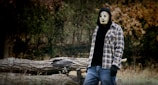 A person wearing a creepy mask stands in an outdoor setting with an axe lodged in a tree trunk in the background. They are dressed in a black hoodie with a checkered shirt over it and blue jeans. The surroundings include some fallen logs and dense trees with autumn-colored foliage.