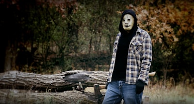 A person wearing a creepy mask stands in an outdoor setting with an axe lodged in a tree trunk in the background. They are dressed in a black hoodie with a checkered shirt over it and blue jeans. The surroundings include some fallen logs and dense trees with autumn-colored foliage.