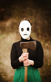 A person wearing a white, homemade-looking mask is holding a large axe. The setting appears to be outdoors, with a blurred, natural background. The individual is dressed in a black top and a green skirt, standing in a straight posture with hands gripping the axe tightly.