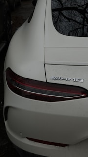 A close-up view of the rear end of a white sports car, displaying the tail light and the AMG emblem. The car's surface is slightly dirty, and the dark surroundings suggest it is parked outdoors, possibly under trees as branches are reflected on the back window.