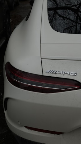 A close-up view of the rear end of a white sports car, displaying the tail light and the AMG emblem. The car's surface is slightly dirty, and the dark surroundings suggest it is parked outdoors, possibly under trees as branches are reflected on the back window.