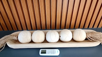 A wooden tray holds five light-colored wooden spheres, each with twine attached on both ends. They are neatly arranged on a dark surface. A small electronic remote control device is placed in front of the tray. Behind, there is a vertically slatted wooden paneling.