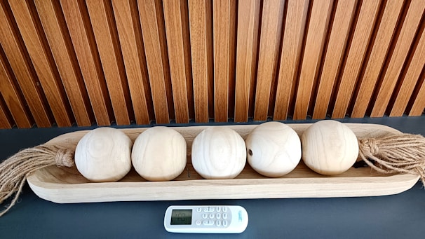 A wooden tray holds five light-colored wooden spheres, each with twine attached on both ends. They are neatly arranged on a dark surface. A small electronic remote control device is placed in front of the tray. Behind, there is a vertically slatted wooden paneling.
