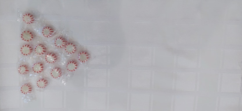 A group of peppermint candies arranged in a triangular pattern on a textured white surface. The candies are individually wrapped and have a red and white striped pattern.