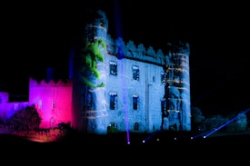 A castle at night illuminated with colorful lights, including vibrant shades of blue and pink. Projections on the castle walls depict a green figure. Various beams of light extend outward creating a dynamic visual effect.