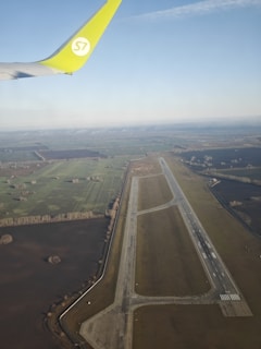 An aerial view of an airport runway and surrounding farmland with a partial view of an airplane wing with a green tip. The landscape shows a mix of agricultural fields and patches of forested areas under a clear sky.