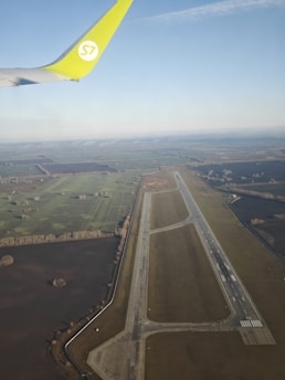 An aerial view of an airport runway and surrounding farmland with a partial view of an airplane wing with a green tip. The landscape shows a mix of agricultural fields and patches of forested areas under a clear sky.
