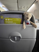 A plush toy bear with a striped shirt and heart emblem is tucked into the back of an airplane seat pocket. The pocket also contains a safety instruction card and magazine with the S7 Airlines logo visible. The aircraft interior is visible, with the overhead bin and lighting in the background.