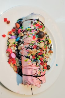 A rolled ice cream dessert topped with colorful sprinkles, nuts, chocolate chips, and small round candies is placed on a white plate. The ice cream has a pink hue, with chocolate syrup drizzled over it and various toppings scattered around, creating a vibrant and indulgent appearance.