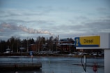 A riverside scene with a gas station sign displaying 'Diesel' in the foreground. Behind the sign, there is a body of water, followed by a line of trees and industrial buildings. Smoke is rising from a chimney, indicating industrial activity. The sky is overcast with clouds.