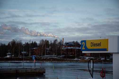 A riverside scene with a gas station sign displaying 'Diesel' in the foreground. Behind the sign, there is a body of water, followed by a line of trees and industrial buildings. Smoke is rising from a chimney, indicating industrial activity. The sky is overcast with clouds.