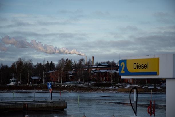 A riverside scene with a gas station sign displaying 'Diesel' in the foreground. Behind the sign, there is a body of water, followed by a line of trees and industrial buildings. Smoke is rising from a chimney, indicating industrial activity. The sky is overcast with clouds.