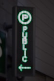 A vertical LED sign with a green digital display shows a letter 'P' enclosed in a circle at the top. Below it, the word 'PUBLIC' is written in vertical alignment. An arrow pointing to the left is seen at the bottom.