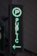A vertical LED sign with a green digital display shows a letter 'P' enclosed in a circle at the top. Below it, the word 'PUBLIC' is written in vertical alignment. An arrow pointing to the left is seen at the bottom.