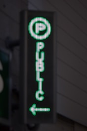 A vertical LED sign with a green digital display shows a letter 'P' enclosed in a circle at the top. Below it, the word 'PUBLIC' is written in vertical alignment. An arrow pointing to the left is seen at the bottom.