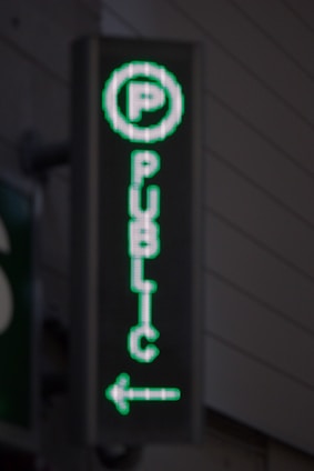 A vertical LED sign with a green digital display shows a letter 'P' enclosed in a circle at the top. Below it, the word 'PUBLIC' is written in vertical alignment. An arrow pointing to the left is seen at the bottom.
