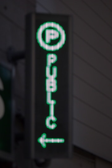 A vertical LED sign with a green digital display shows a letter 'P' enclosed in a circle at the top. Below it, the word 'PUBLIC' is written in vertical alignment. An arrow pointing to the left is seen at the bottom.