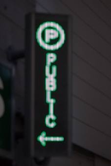 A vertical LED sign with a green digital display shows a letter 'P' enclosed in a circle at the top. Below it, the word 'PUBLIC' is written in vertical alignment. An arrow pointing to the left is seen at the bottom.