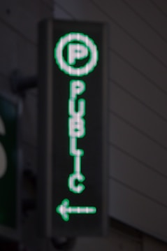 A vertical LED sign with a green digital display shows a letter 'P' enclosed in a circle at the top. Below it, the word 'PUBLIC' is written in vertical alignment. An arrow pointing to the left is seen at the bottom.