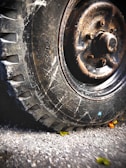 A close-up of a rugged, worn tire with visible treads and bolts on a metallic hub. The surface has signs of rust and wear, indicating age and use. The tire rests on a rough, textured road, adorned with scattered small leaves, both green and yellow, that add a slight contrast to the earthy tones.