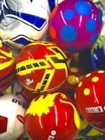 A collection of colorful balls with various patterns and designs, including stripes, polka dots, and geometric shapes. The balls have vibrant colors like red, yellow, purple, and blue. The surface of each ball is glossy and reflective.