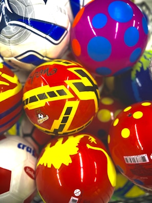 A collection of colorful balls with various patterns and designs, including stripes, polka dots, and geometric shapes. The balls have vibrant colors like red, yellow, purple, and blue. The surface of each ball is glossy and reflective.
