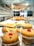A display of assorted pastries on a glass shelf, featuring two small cakes with cherries on top in the foreground. Behind them, various pastries including croissants are arranged, with a blurred background of the bakery setting.