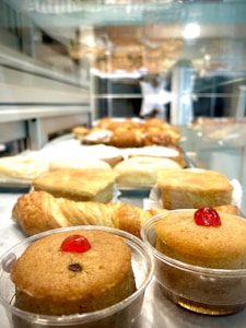 A display of assorted pastries on a glass shelf, featuring two small cakes with cherries on top in the foreground. Behind them, various pastries including croissants are arranged, with a blurred background of the bakery setting.