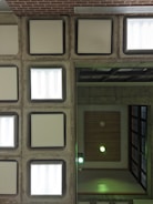 An architectural ceiling view with symmetrical, square light fixtures set in a grid pattern. Concrete frames with white, frosted light panels dominate one half, while the other half features wooden paneling with recessed lighting emitting a greenish glow.