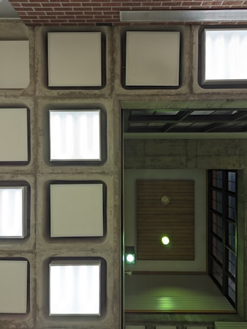 An architectural ceiling view with symmetrical, square light fixtures set in a grid pattern. Concrete frames with white, frosted light panels dominate one half, while the other half features wooden paneling with recessed lighting emitting a greenish glow.