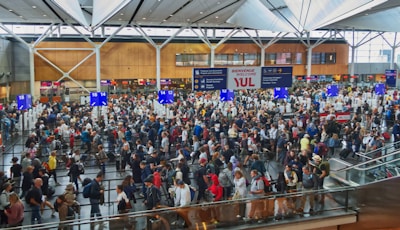 A bustling airport terminal filled with a large crowd of people queuing at check-in desks. The high ceiling and modern architecture are visible, along with various signs and electronic displays. The scene is lively, with people of different ages and backgrounds moving in different directions.