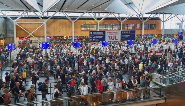 A bustling airport terminal filled with a large crowd of people queuing at check-in desks. The high ceiling and modern architecture are visible, along with various signs and electronic displays. The scene is lively, with people of different ages and backgrounds moving in different directions.
