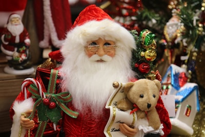 A festive Santa Claus figurine is surrounded by various Christmas decorations. The figure is holding a teddy bear and a scroll in one hand, and a wrapped gift with a candy cane in the other. The background includes more Christmas-themed items like a small house ornament and additional Santa figurines. The scene is rich with holiday colors and textures, emphasizing a warm and celebratory atmosphere.