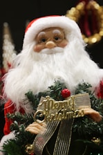 A festive decoration featuring a Santa Claus doll with a white beard and red hat, holding a small Christmas tree adorned with a golden ribbon and the words 'Merry Christmas' in gold lettering. The background includes blurred holiday ornaments.