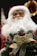A festive decoration featuring a Santa Claus doll with a white beard and red hat, holding a small Christmas tree adorned with a golden ribbon and the words 'Merry Christmas' in gold lettering. The background includes blurred holiday ornaments.