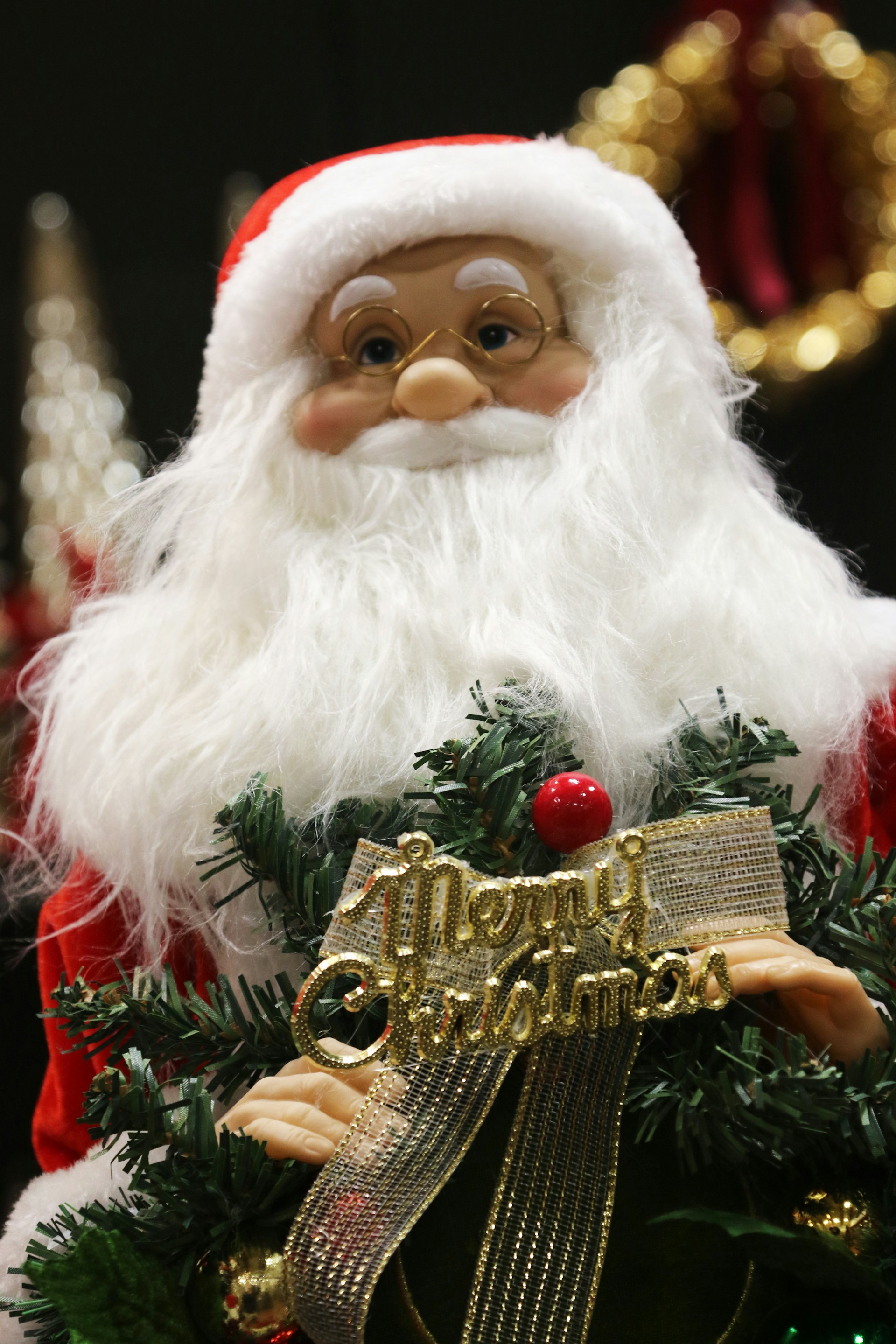 A festive decoration featuring a Santa Claus doll with a white beard and red hat, holding a small Christmas tree adorned with a golden ribbon and the words 'Merry Christmas' in gold lettering. The background includes blurred holiday ornaments.