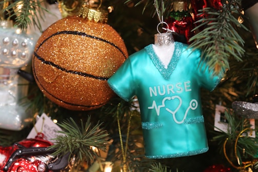 A festive Christmas tree decorated with a nurse-themed ornament shaped like a teal scrub top with medical symbols, alongside a glittering gold basketball ornament. The ornaments are surrounded by green pine branches with other decorations peeking through.