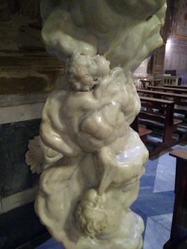 A detailed marble sculpture depicting intertwined figures in a dramatic pose, located inside a richly decorated building. The craftsmanship focuses on intricate detailing of human anatomy and expressions. Background elements include wooden furniture and elaborate wall decorations.