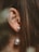 A close-up of an ear adorned with a pearl earring, set against long, brown hair. The earring features two pearls, one smaller and one larger, connected by a thin chain.