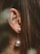 A close-up of an ear adorned with a pearl earring, set against long, brown hair. The earring features two pearls, one smaller and one larger, connected by a thin chain.