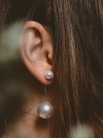 A close-up of an ear adorned with a pearl earring, set against long, brown hair. The earring features two pearls, one smaller and one larger, connected by a thin chain.