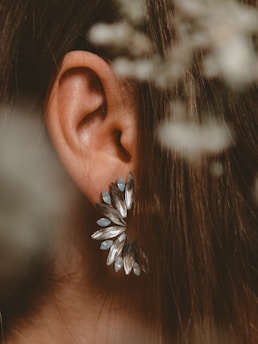 A close-up of a person's ear adorned with a large, decorative earring. The earring is designed with multiple elongated metallic and bluish stones arranged in a fan-like pattern. Strands of brown hair frame the ear, and there is a soft-focus foliage in the foreground.