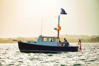 A small motorboat with a cabin is floating on a body of water. The boat has several people on board and is equipped with two flags, one of which is the American flag. The atmosphere is serene with a slight haze, and the background shows a blurred outline of distant trees or land.