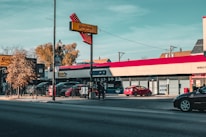 A commercial street scene with several storefronts and cars parked along the road. A prominent red Giordano's sign is visible, with a clear sky and a few trees in the background. Signs indicate some businesses, such as a Geico office and a pizza restaurant.