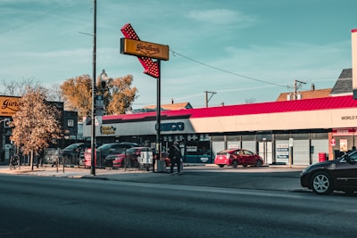 A commercial street scene with several storefronts and cars parked along the road. A prominent red Giordano's sign is visible, with a clear sky and a few trees in the background. Signs indicate some businesses, such as a Geico office and a pizza restaurant.