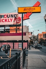 A street scene captures a Giordano's sign with bright red and yellow colors, featuring an illuminated arrow. A large advertisement billboard is visible on the left, displaying a man in a red shirt holding a basketball, partially obscured by the sign. Below the billboard, a woman stands in a parking lot holding shopping bags with a smile, dressed in dark clothing. On the right side of the image, a person with a suitcase walks down the sidewalk beside a street with visible cars. A 'For Lease' sign is posted on the building in the background.