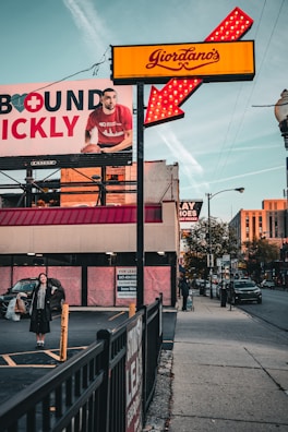 A street scene captures a Giordano's sign with bright red and yellow colors, featuring an illuminated arrow. A large advertisement billboard is visible on the left, displaying a man in a red shirt holding a basketball, partially obscured by the sign. Below the billboard, a woman stands in a parking lot holding shopping bags with a smile, dressed in dark clothing. On the right side of the image, a person with a suitcase walks down the sidewalk beside a street with visible cars. A 'For Lease' sign is posted on the building in the background.